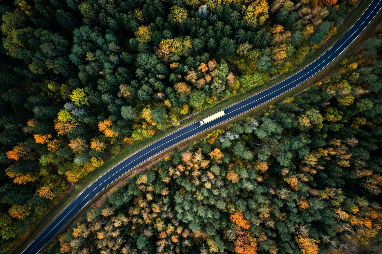Aerial view of a commercial truck driving along a highway through forested terrain, highlighting long-haul route planning to reduce toll road costs.